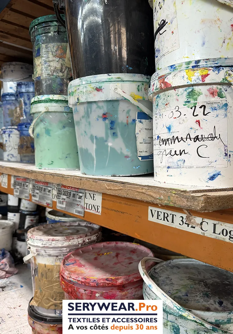 Pots d'encres de sérigraphie colorées rangés sur des étagères dans un atelier de marquage textile.