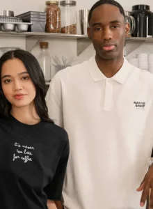 Deux personnes portent un t-shirt noir et un polo blanc Native Spirit dans un espace de café ou de restauration.