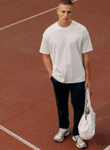 Personne portant un t-shirt blanc Native Spirit avec un pantalon noir et un sac en toile blanc sur une piste de sport.